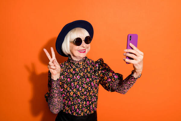 Photo of smiling cheerful positive good mood old woman take selfie on phone showing v-sign isolated on orange color background